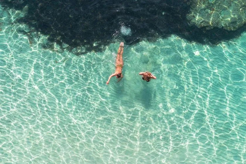 Two swimmers seen from above in a clear turquoise cove, sunlight rippling across the sand below